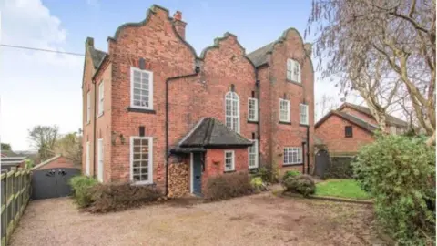 Claire Massey  A view of the front of Claire Massey's house from the drive before the fire in February 2023. We can see 10 windows in the brick wall of the facing wall of the home with bushes around the front and a gravel drive