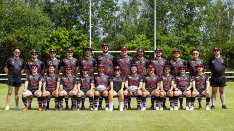 A rugby team is pictured on a pitch for an official picture. There are two lines of players - one line is sitting and the other is standing. They are all smiling at the camera, with the rugby posts visible behind them. They are all wearing branded black and pink sunhats and their full kit, which is black with pink and multi-coloured patterns on it. 