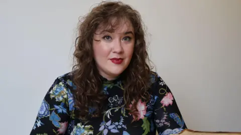 University College, University of Oxford Dr Leah Veronese is wearing a black blue and pink top. She has long curly brown hair and is in front of a white wall.