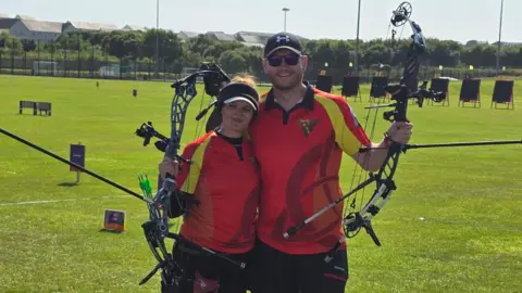 ISLE OF MAN ARCHERY TEAM Aalin George and Ethan Moore wearing team Isle of Man red and yellow tops and holding up their archery bows. The archery field with its targets and markers is laid out on grass behind them.