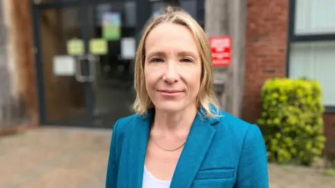 A woman with shoulder length blonde hair, wearing a blue suit jacket and white top.