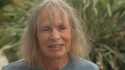 Jamie Niblock/BBC A head and shoulders image of a woman wearing a light blue top. She is looking just past the camera and standing in front of some plants.
