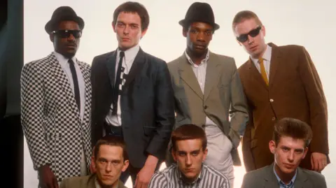 An archive photo showing seven men in a group, with four standing and three sitting or kneeling at the front. Most of them are wearing suit jackets and skinny ties.