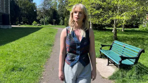 Jess wearing a blue Aztec-design waistcoat and blue and white striped trousers. She is walking in a park with a bench to her right and grass and trees behind her.