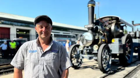 Alex Sharphouse is wearing a short sleeved grey shirt and flat cap and is smiling standing next to a large steam locomotive on the right. It is a sunny day.