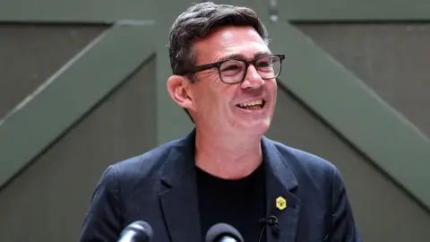 Andy Burnham smiling and wearing a black suit in front of a green door