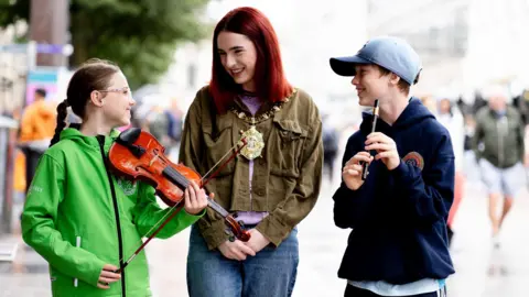 Belfast City Council A girls in green jacket holding violin, a woman with dark green jacket, mayoral chains and denim jeans and a boy with navy jumper and blue hat holding a flute