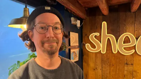 Peter Hefferman smiles for a camera inside his cafe. He is wearing a black beanie hat and round-rimmed glasses with a goatee beard. In the background is the logo of his cafe with a pot plant on a shef. 