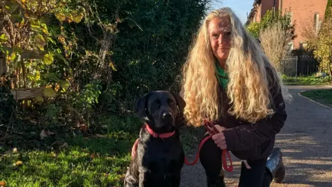 A woman with long, curly blond hair holds her black Labrador dog by a red lead. They are crouching on a footpath by green grass and shrubs.