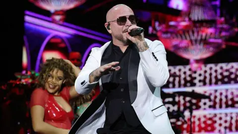 Pitbull performs onstage. He is a bald man wearing shades and a white suit, holding a microphone. A female backing dancer wearing a red dress is dancing behind him.