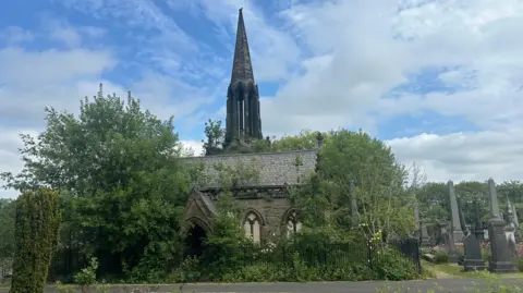 Jessica Bayley/ BBC An outside view of a 19th Century chapel with alongside trees and some tall gravestones on a clear day.
