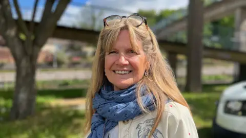 A woman with long light brown hair, glasses positioned up on her head and a blue spotted scarf wrapped around her neck smiling and standing in front of the blurred background of a footbridge over a highway.