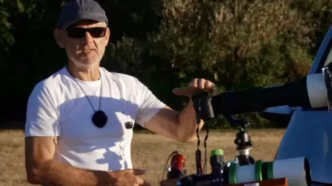 Simon Wallace/Simon's Astro A man in a white T-shirt, and wearing a grey baseball cap and sunglasses, rests his left hand on a telescope. He is standing on a grassy area with hedging behind him. 