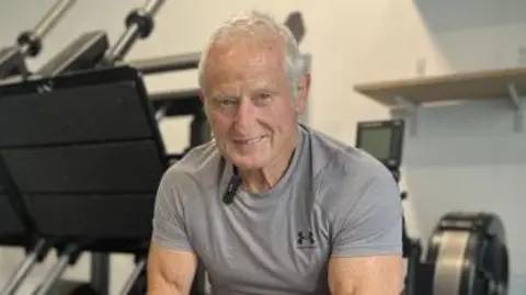 University of Wales Trinity Saint David Peter is wearing the a grey gym t-shirt, and is smiling for the camera as he sits on a weight lifting bench. There are exercise machines behind him and to the left of him.