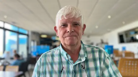A man in a green and white checkered shirt with grey hair smiling at the camera.Guerns