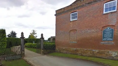 Red brick wing of Fawsley Hall with stone gateposts and hedge-lined drive