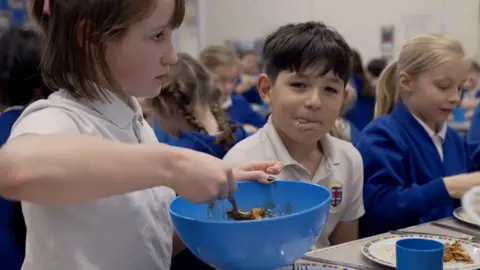Three children sit at a dining hall table in a school canteen, one boy with brown hair is licking his lips, another girl is spooning food out of a blue bowl.