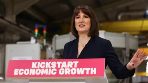 Rachel Reeves is smartly dressed with tidy dark-brown shoulder-length hair and a fringe across her forehead. She wears a dark blue-black blazer with a V-neck. She seems to be mid-sentence, and is gesticulating slightly, holding her left hand up at her side. She is stood behind a podium which says "kickstart economic growth" in capital white lettering on a red background.