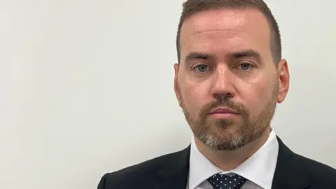 A man with short, light brown hair, wears a blue suit, white shirt and blue polka dot tie. He has a short bear and stands against a plain white background with a neutral expression on his face.
