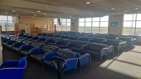 Isle of Man Airport A large number of blue leather seats in the rear section of the departures lounge, large windows are on the back right wall.