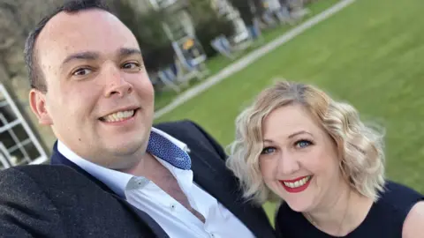 Family handout Laurent and his wife Gemma are standing close together outdoors, smiling for a selfie. Behind them is a green lawn with several lounge chairs and a modern building with large windows. Gemma, on the right, has short wavy blonde hair and wears a dark top; Laurent wears a dark jacket over a white shirt with a patterned collar.

