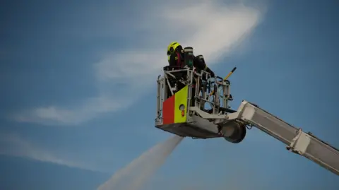 Two fire officers stand on an aerial platform from which water is being poured. The blue sky can be seen behind them.