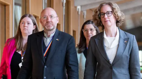 PA Media Scottish Green Party co-leaders Patrick Harvie (second left) and Lorna Slater (right) walking in the Scottish Parliament, with two women walking behind them. Mr Harvie is wearing a three-piece blue suit and an open necked shirt, while Ms Slater is wearing a grey suit jacket and a cream top.