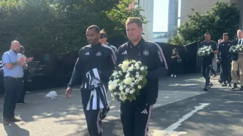 Newcastle United head coach Eddie Howe walks with club players and staff carrying a floral wreath of white and cream flowers with green foliage for Kevin Keegan at St James' Park. Several bystanders clap as he walks by.