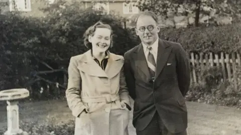 Marcia Lewis A black and white photo of a Elva and Anthony Pratt on a lawn. There is a bird bath behind them to the left and a hedge beyond that. Anthony Pratt is wearing dark-rimmed circular glasses and a suit. His wife (to the left is in a light coloured coat
