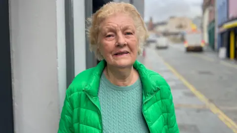 Veronica Doherty is standing in a street. She's wearing a green coat and a light green jumper. She has short hair.