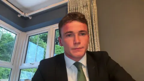 A young man with a black suit, green tie and white shirt. he has short brown hair and stares into the camera. 