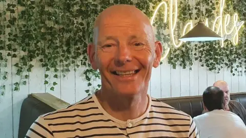 A smiling Declan Moore stands in his cafe in Derry. Two customers are seated at a table to his right. He is wearing a black and white striped t-shirt. Ivy hangs above a row of seats in the background of the picture.