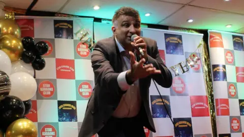 Star Karaoke Club A man in a black suit jacket and white shirt is singing into a microphone and has his arm outstretched. Behind hi are gold, black and white balloons.