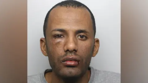 A mugshot of Ibrahim Albashir. He has short, dark hair and brown eyes. He has a cut to his lip and a bruise beneath his right eye. He is wearing a grey jumper.