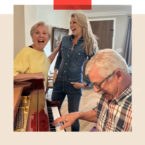 A smiling Marijeke stands next to Lara Levington while Marijeke's husband Tom plays the piano.