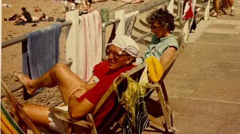 An old picture of Colin and Doreen Hunt sunbathing in deckchairs on the promenade. The photo is orange-tinted, there are towels hanging over railings, and sand in the background