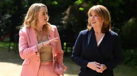 Rachel Reeves on the right, smiling in a blue suit, being interviewed by Laura Kuenssberg on the left, in a pink suit, in a garden as the two of them walk side by side