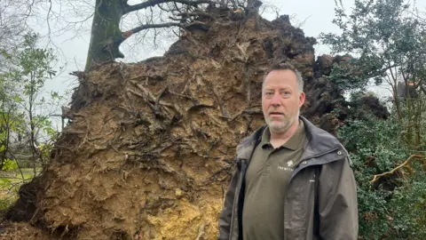Gary Long stands in front of a huge root ball of a tree which has fallen. He looks serious. He has short brown and grey hair and a grey beard. He is wearing a Trewithen-branded top under a wax jacket.