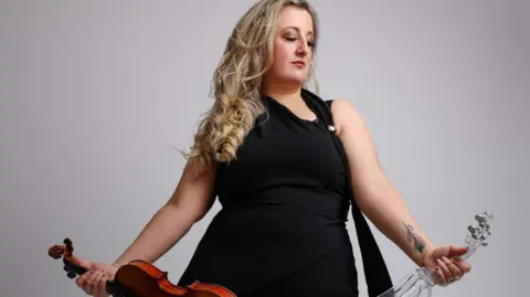 Laura Macmillan Image shows Laura Macmillan, a classically trained musician. She is holding a violin in each hand. Laura is wearing a black dress and has long blonde hair. 