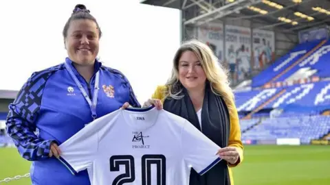 Tranmere Rovers FC Hannah Naylor wears a Tranmere top and dark trousers, while Jessica Gallier Booth stands next to her at the Tranmere ground. Ms Gallier Booth is wearing a mustard-coloured jacket, white top and black scarf. They are holding a Tranmere Rovers women's team shirt with the number 20 and The Martin Gallier Project logo on the back. 