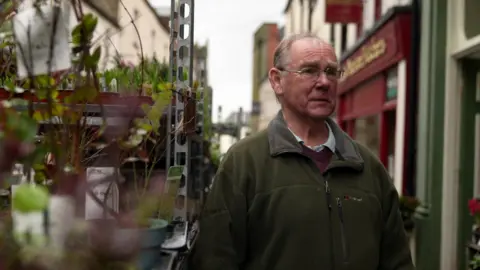 John Meynell stands by an outdoor display of plants. He is an older man with short grey hair, silver-rimmed glasses, a green fleece and a blue shirt.