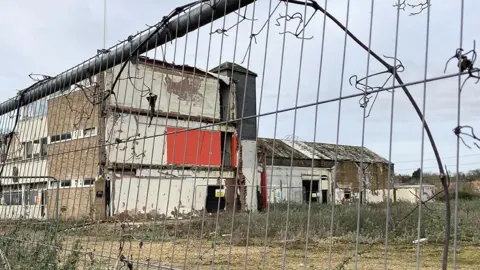 A view of a derelict factory through a silver metal fence. The building is partially demolished with broken windows and is surrounded by overgrown grass and weeds.
