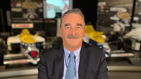 A man in the foreground torso up wearing a suit and tie, with brown hair and a brown moustache. In the background and out of focus is a formula one car