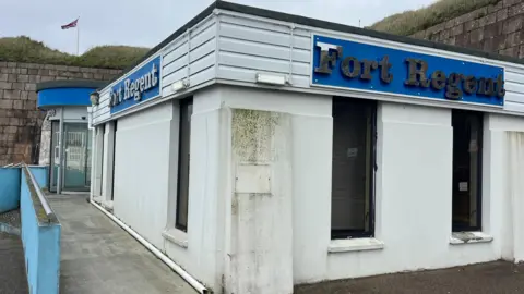 BBC Exterior of a small white building with blue “Fort Regent” signs, a concrete walkway, and a Union Jack flag on a grassy embankment behind.
