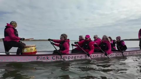 Pink Champagne boat with two single file lines rowing whilst a woman at the end bangs a drum.