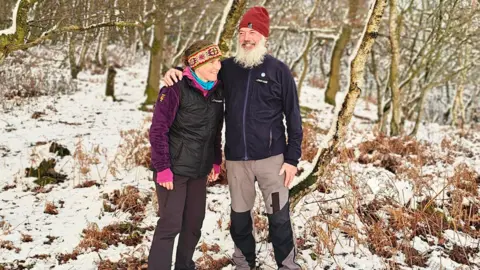 Zoe Langley-Wathen and her husband Mike Langley. They are stood in a snowy forest. Zoe, left, has on a colourful red, green and white head warmer, and a black and purple coat. Mike has his arm around her and is wearing a dark blue fleece with a purple zip, along with a dark red beanie hat.
