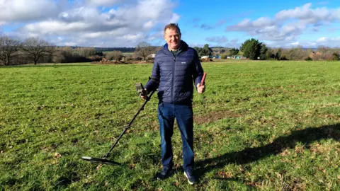 A man stands in a field on a sunny day. He is holding a metal detector. 