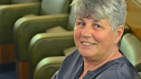 Councillor Yvonne Gagen with short grey hair wearing a grey top. She is sitting in the council chamber with empty green leather seats in the background. She is smiling.