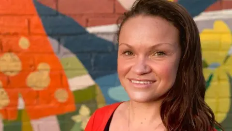 A woman with brown hair. She is standing smiling at the camera. She is stood in front of a multi-coloured mural painted on a brick wall.