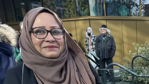 Shantona Women's and Family Services A close-up of Nahid Rasool, who wears a brown hijab and rounded black glasses. She is smiling slightly.